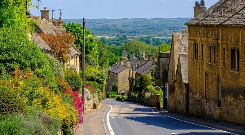 A flower-lined Cotswolds village street curving toward distant countryside on a combo day trip
