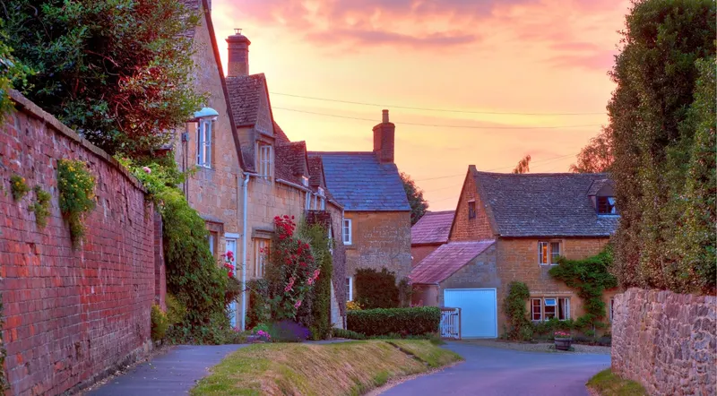 A quiet Cotswolds village lane of honey-stone cottages under a pink evening sky